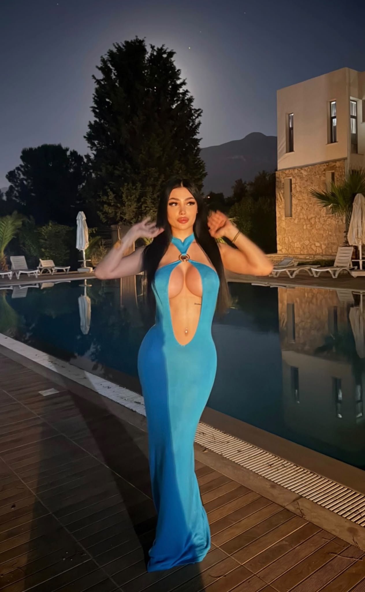 Woman in a bright blue cutout gown standing by a pool at night, moonlit backdrop and resort buildings in the background.
