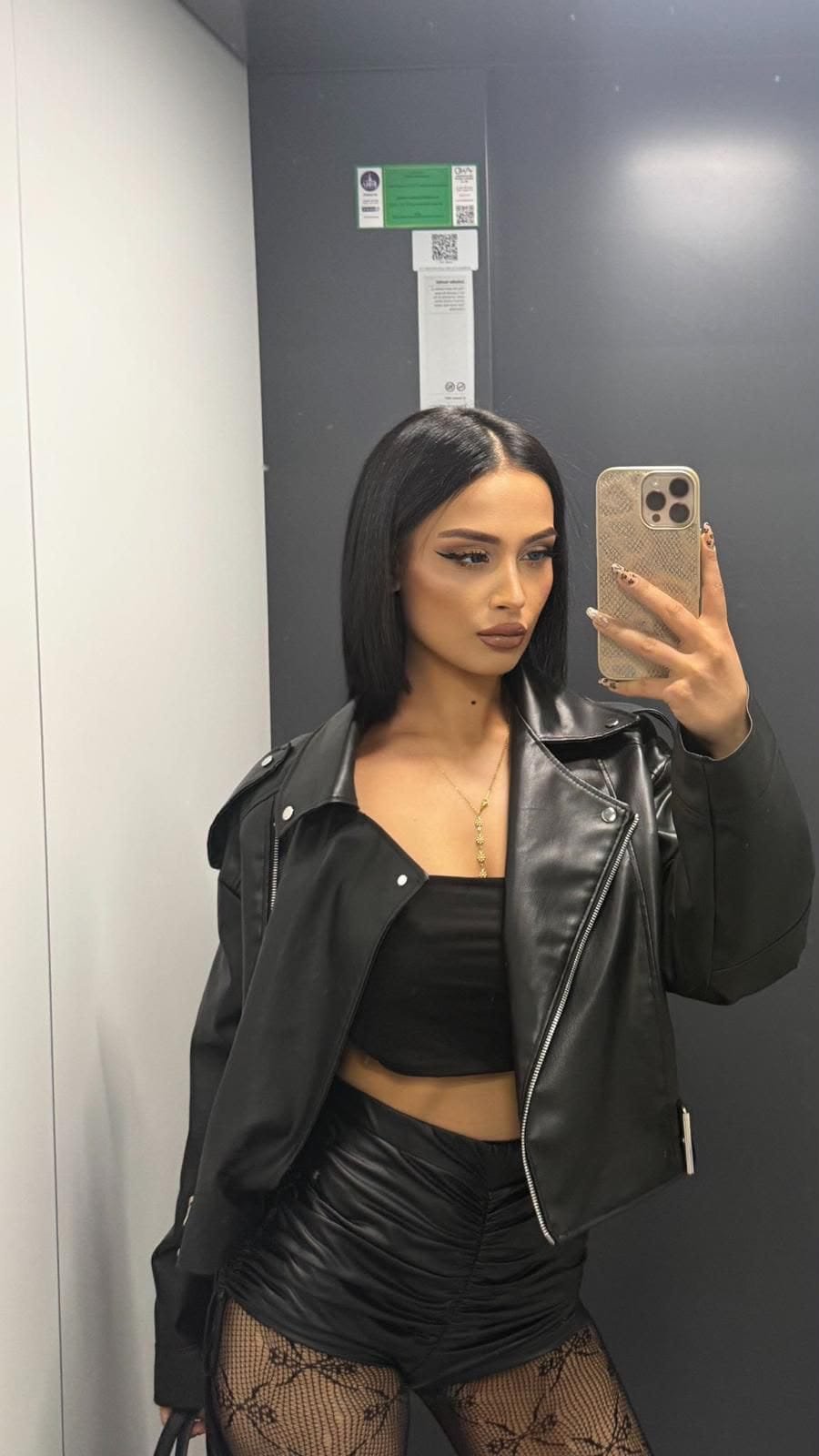 Woman in a black leather jacket, crop top, and ruched shorts posing for a mirror selfie in an elevator; fishnet tights and gold necklace visible