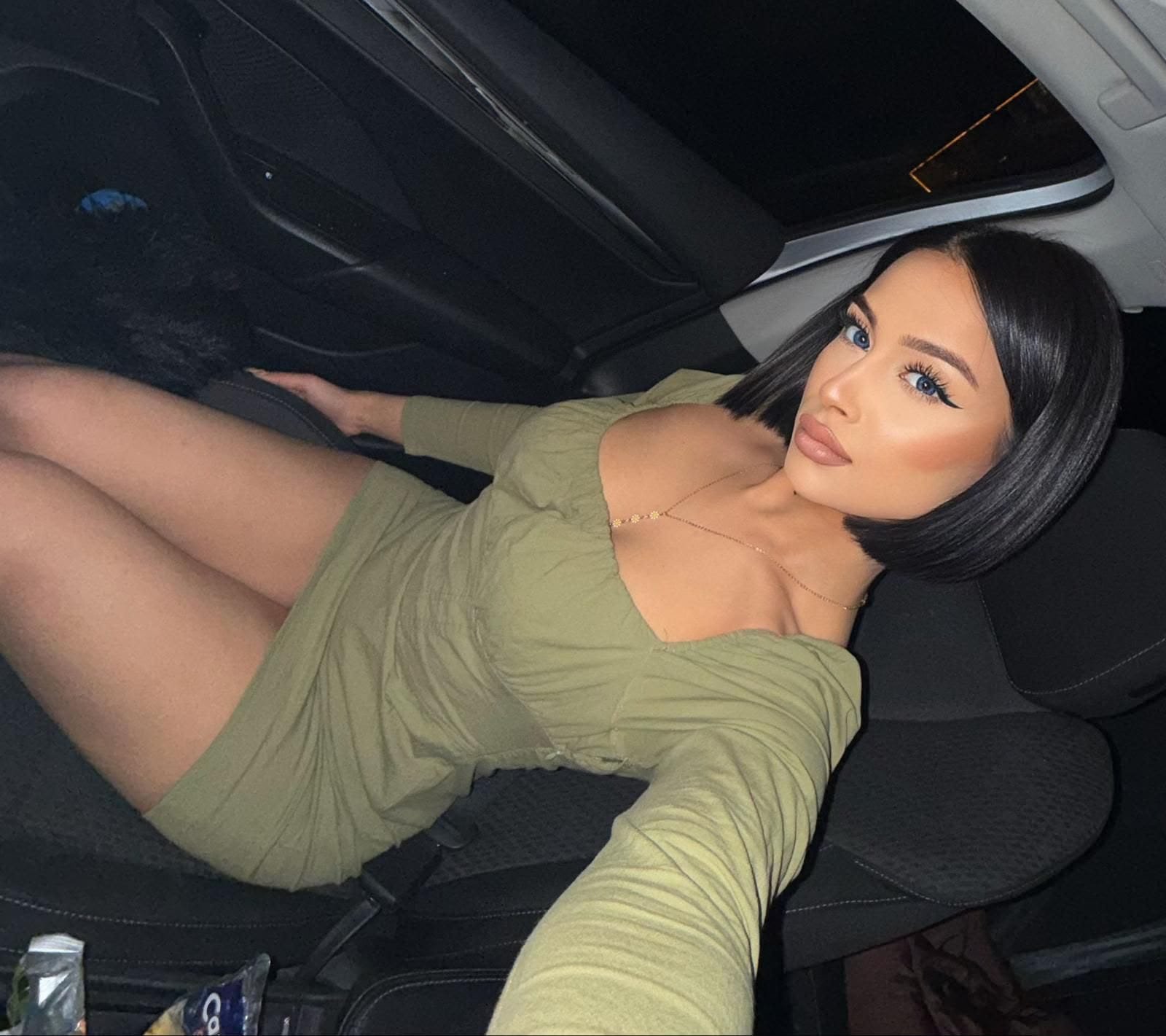 Woman with short black hair and bold makeup sits in a car, taking a selfie in a green dress.