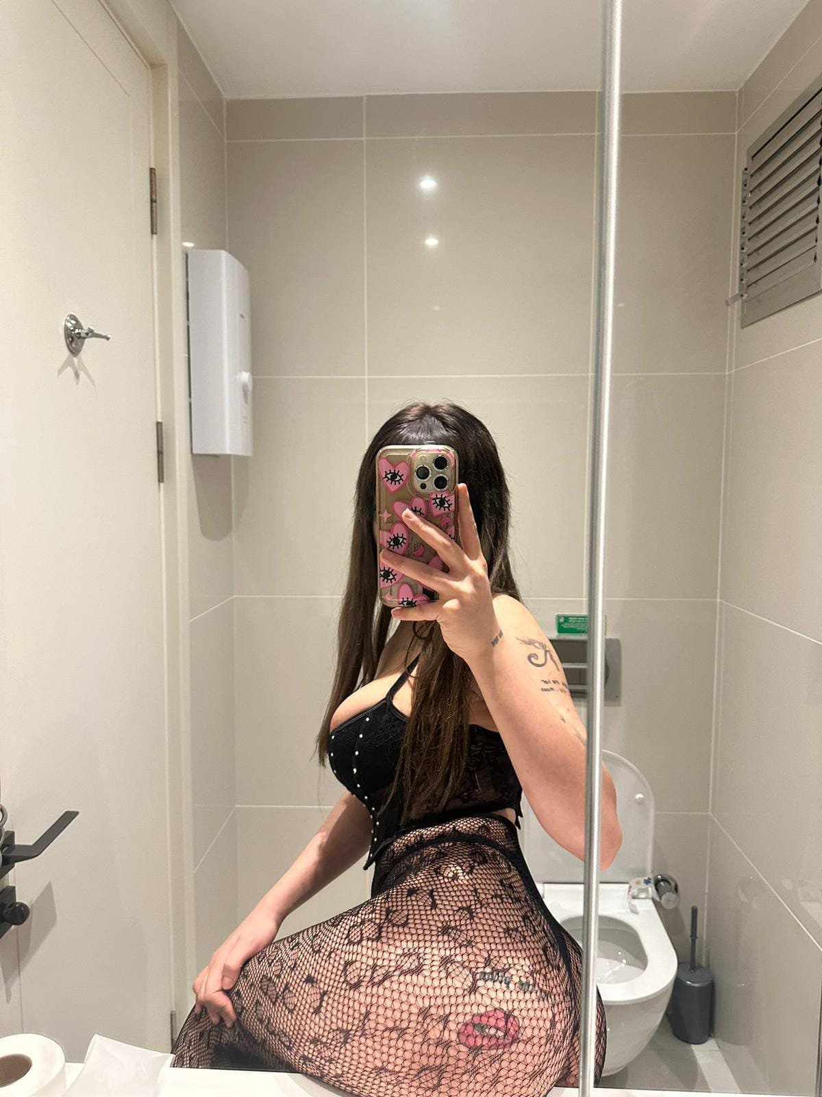 Person taking a mirror selfie in a bathroom, wearing black lingerie and fishnet stockings, phone with a pink case visible in the shot.