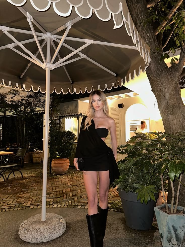 Woman in a black one-shoulder mini dress and tall boots posing under a large outdoor umbrella at night on a cafe patio.