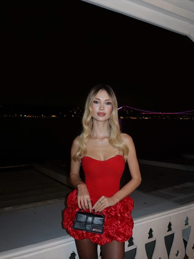 Woman in a red strapless dress standing on a balcony at night, holding a small black handbag with a city lights backdrop and a bridge in the distance beneaththe night sky.