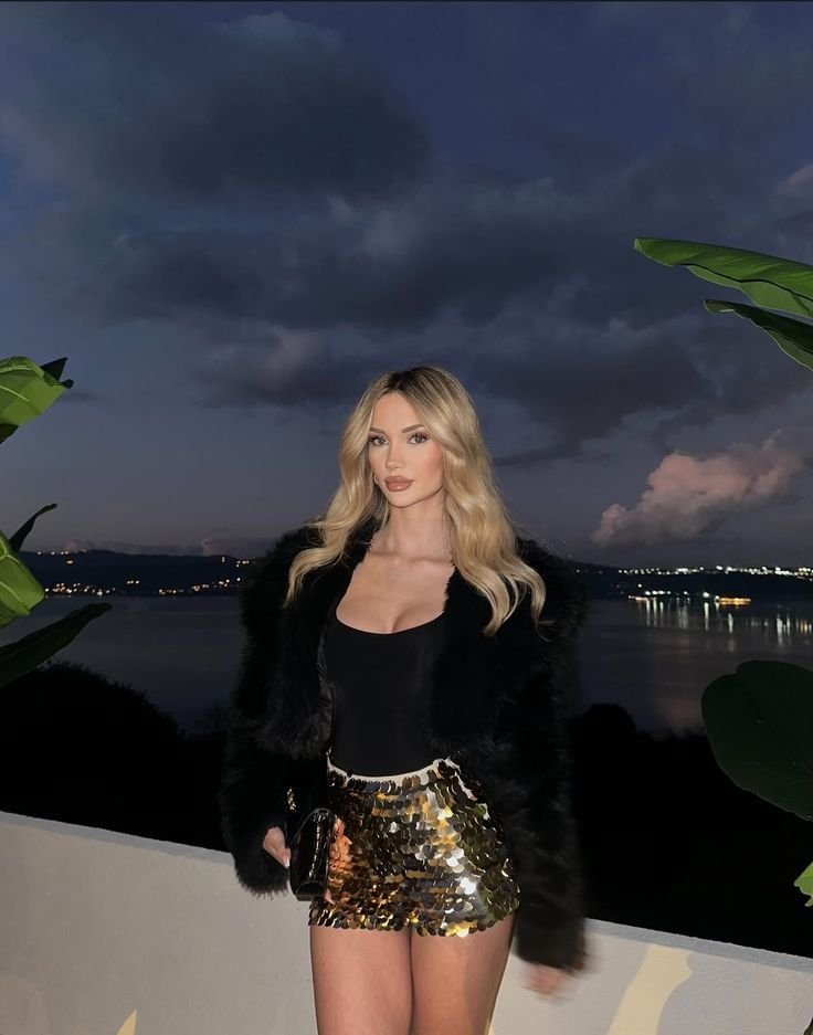 Woman with long blonde waves posing by a railing at dusk, wearing a black top, black fur jacket, and a gold sequined mini skirt by the water edge.