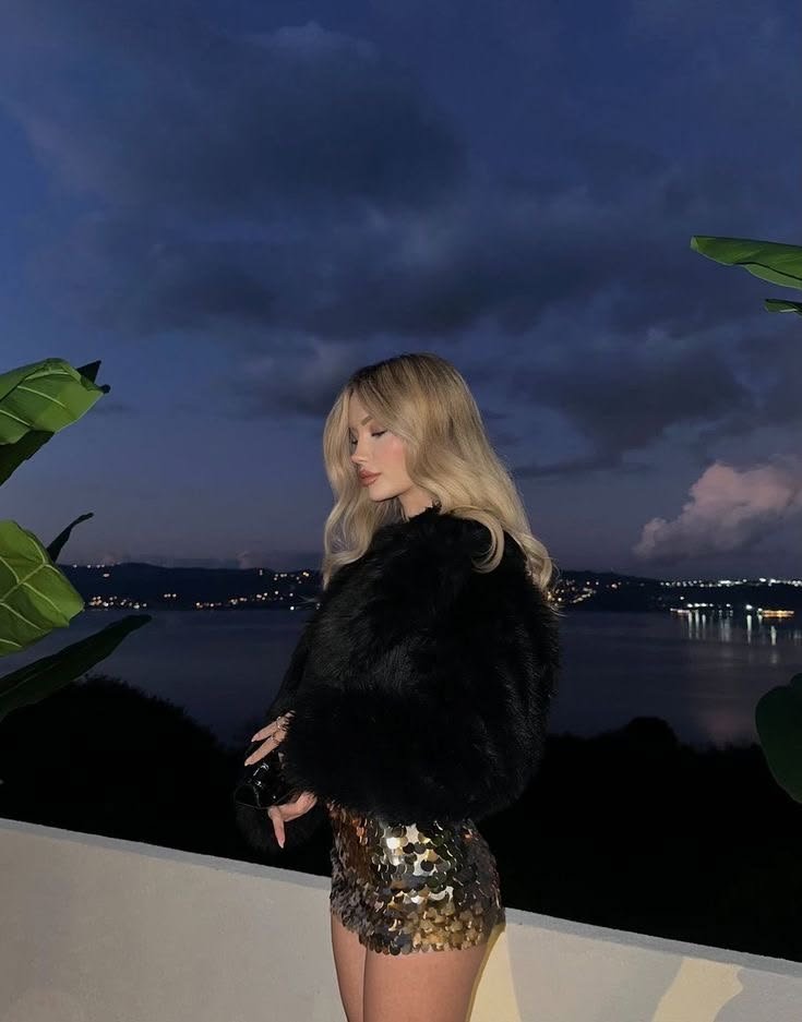 Side-profile of a fashion-forward woman in a black fur coat and gold sequin mini skirt, posing on a balcony by a dark waterway at dusk with distant city lights behind her.