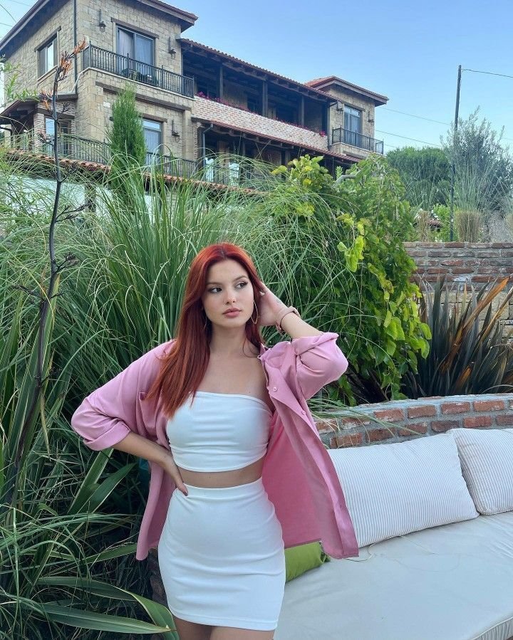 Woman with red hair posing outdoors in a pink button-down shirt over a white tube top and mini skirt, standing by tall grasses and a stone house backdrop.