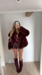 Person posing in a burgundy fur coat, matching top, leather shorts, and knee-high burgundy boots, taking a mirror selfie in a minimalist room.