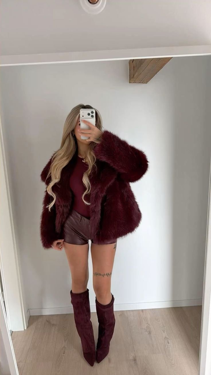 Person posing in a burgundy fur coat, matching top, leather shorts, and knee-high burgundy boots, taking a mirror selfie in a minimalist room.