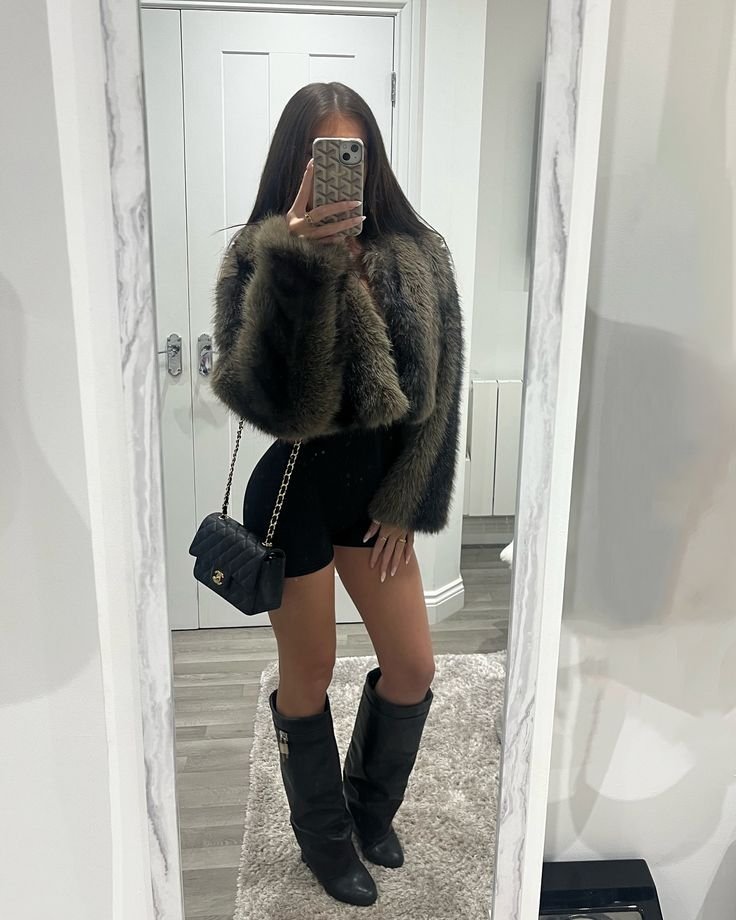 Person taking a mirror selfie in a fur coat, black shorts, knee-high boots, and a small black quilted purse with chain strap.