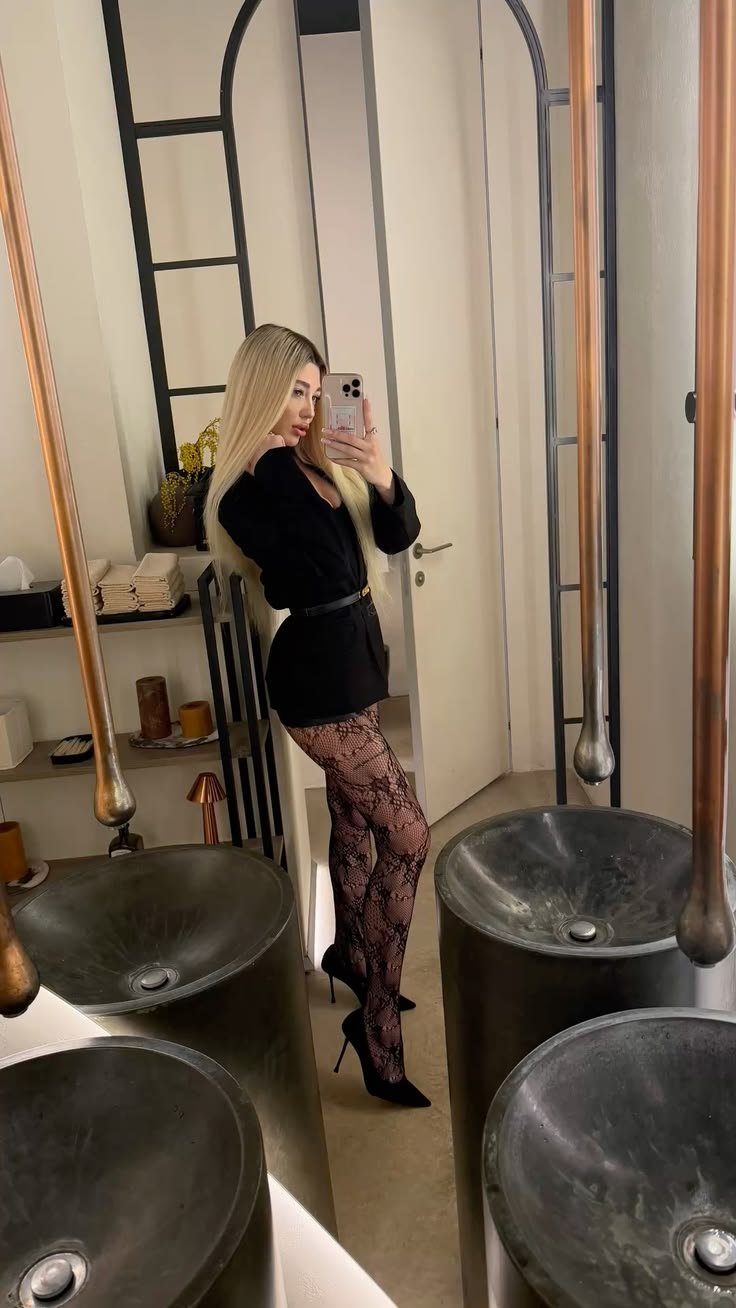 Woman in a black belted blazer dress with lace tights and black heels posing for a mirror selfie in a room with industrial-style fixtures and basins.