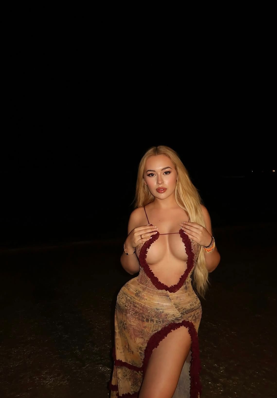 Young woman with long blonde hair posing at night outdoors, wearing a patterned dress with burgundy trim and a high slit.