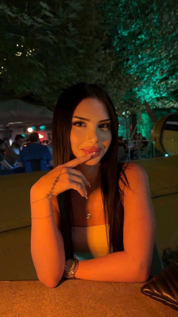 Young woman with long dark hair sits at an outdoor night venue, touching her lips with her finger.” (informative portrait)
