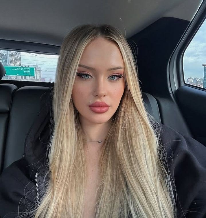 Young woman with long blonde hair and pink makeup looking at the camera while seated in the back of a car, city skyline visible through the window behind her.