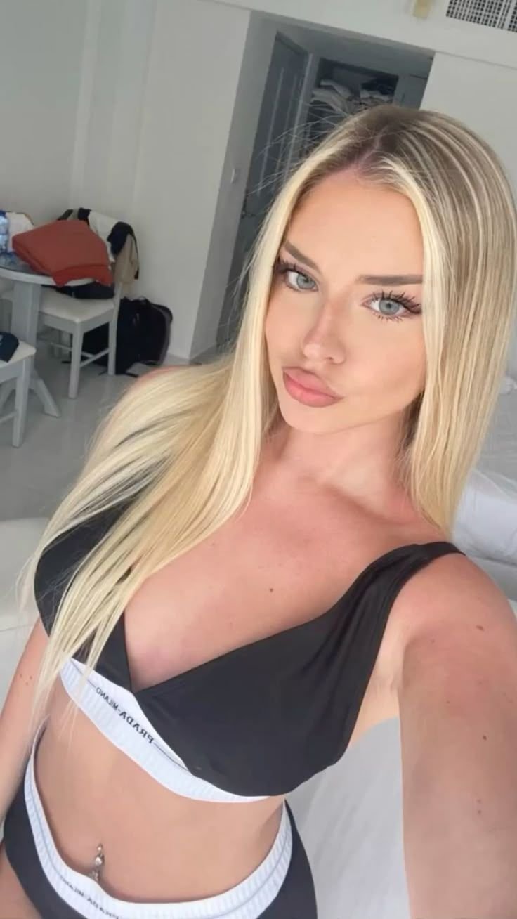 Selfie of a young woman with long blonde hair wearing a black bikini top with white trim, posing in a bright room.