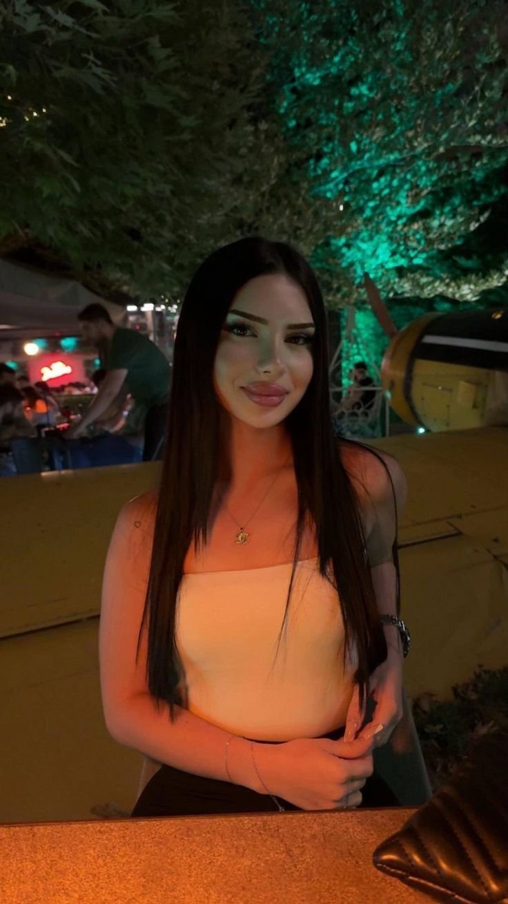 Young woman with long dark hair and a white strapless top sits at a warmly lit outdoor bar at night.