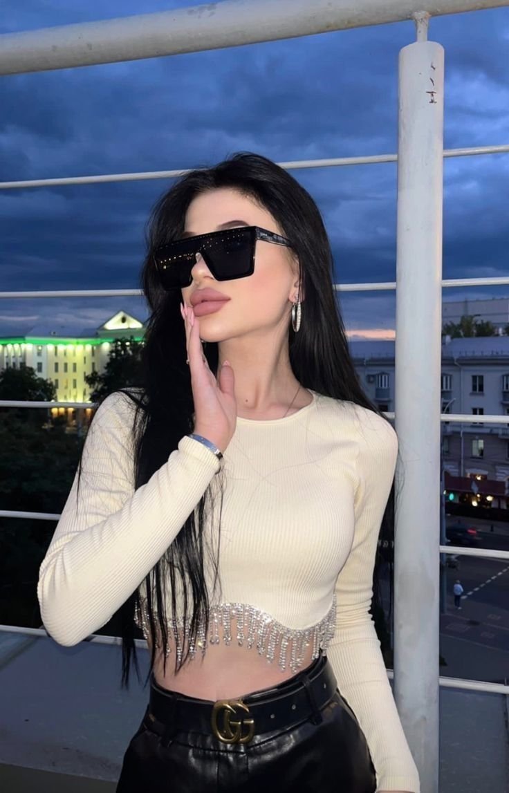 Fashionable woman posing on a rooftop balcony in a cream cropped sweater with jeweled fringe, black sunglasses, and hoop earrings at dusk.