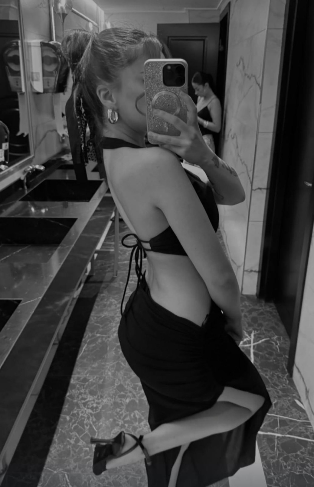 Woman in a backless black dress and high heels takes a mirror selfie in a marble-floored bathroom, phone covering her face.