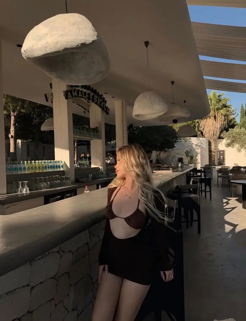Woman in a brown bikini top and black cover-up leaning on an outdoor bar counter at a resort, with large pendant lamps overhead and palm trees in the background.