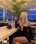 Woman with blonde hair sits at a marble bar counter in a black cut-out dress, palm plant behind her, city lights in the evening.