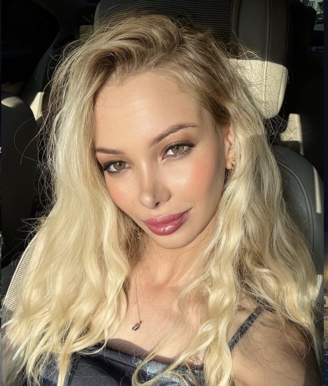 Close-up portrait of a blonde woman with wavy hair in a car, wearing makeup and a delicate necklace.