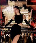 Woman in a black crop top and long skirt posing with eyes closed against a railing in a chandelier-lit bar setting.