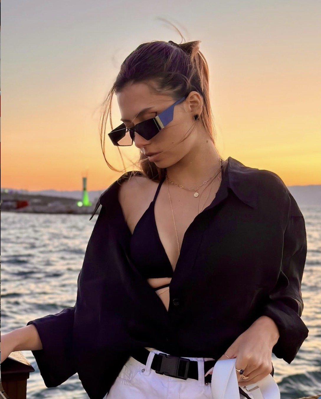 Woman in a black top and white pants by the water at sunset, wearing sunglasses and gold layered necklaces.