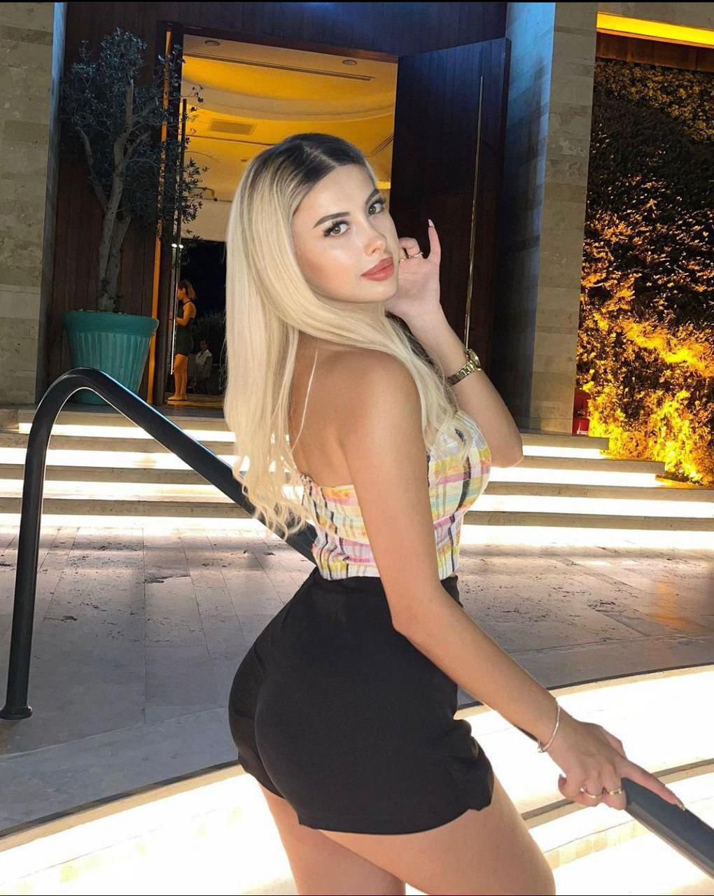 Woman with long blonde hair in a colorful plaid top and black shorts posing on lit outdoor stairs at night.