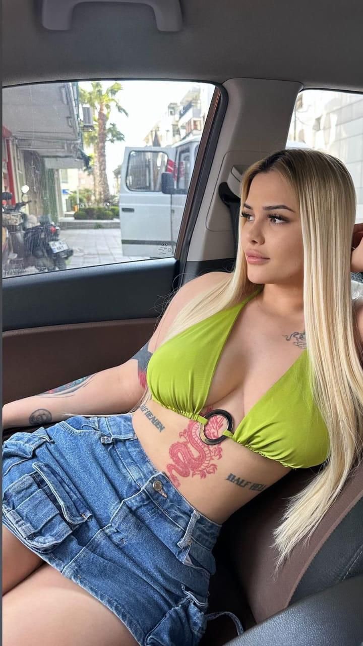 Young woman with long blonde hair wearing a lime green bikini top and denim skirt sits in a car, looking out the passenger-side window with tattoos visible on her arms and torso.