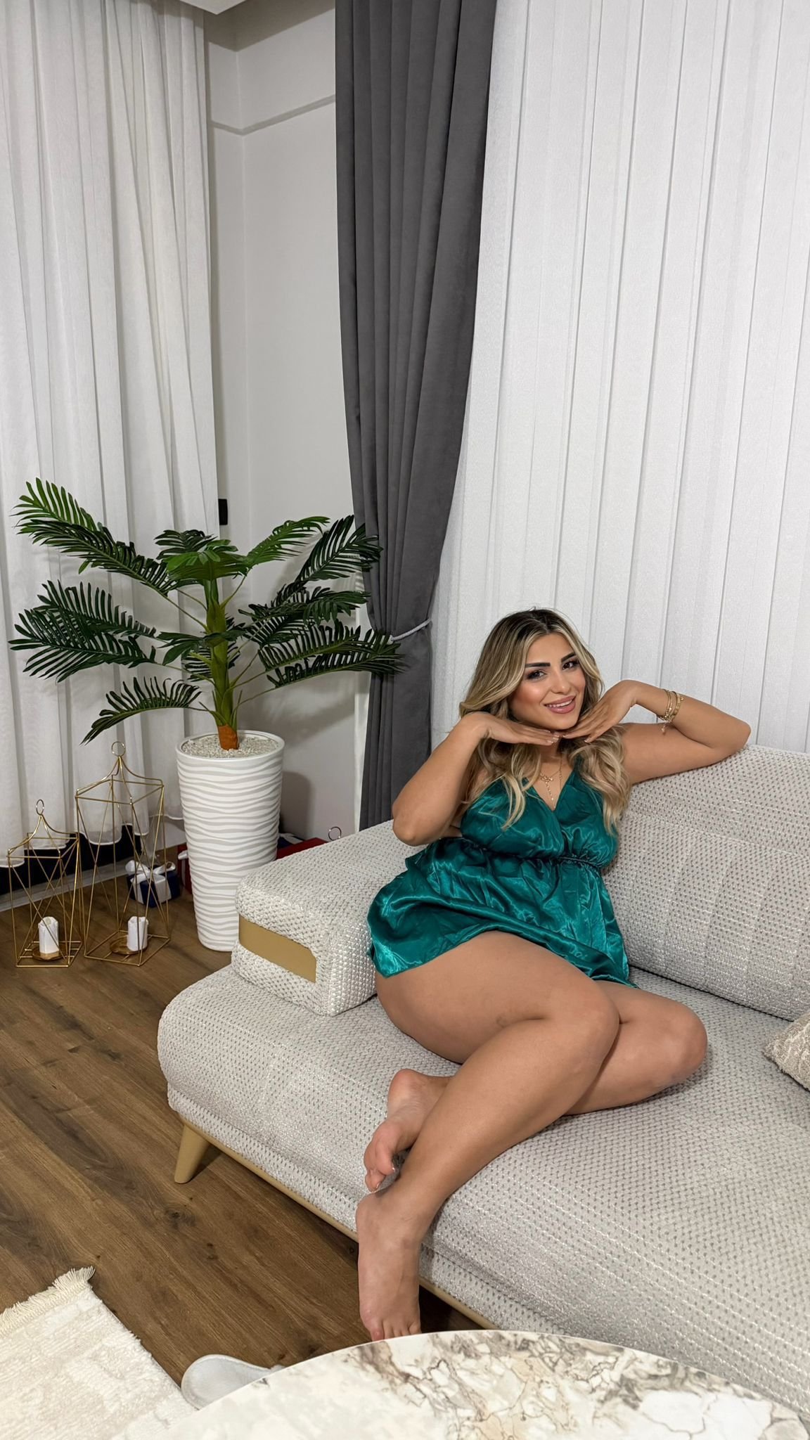 Woman in a teal dress posing on a beige sofa indoors, hands under chin, smiling.
