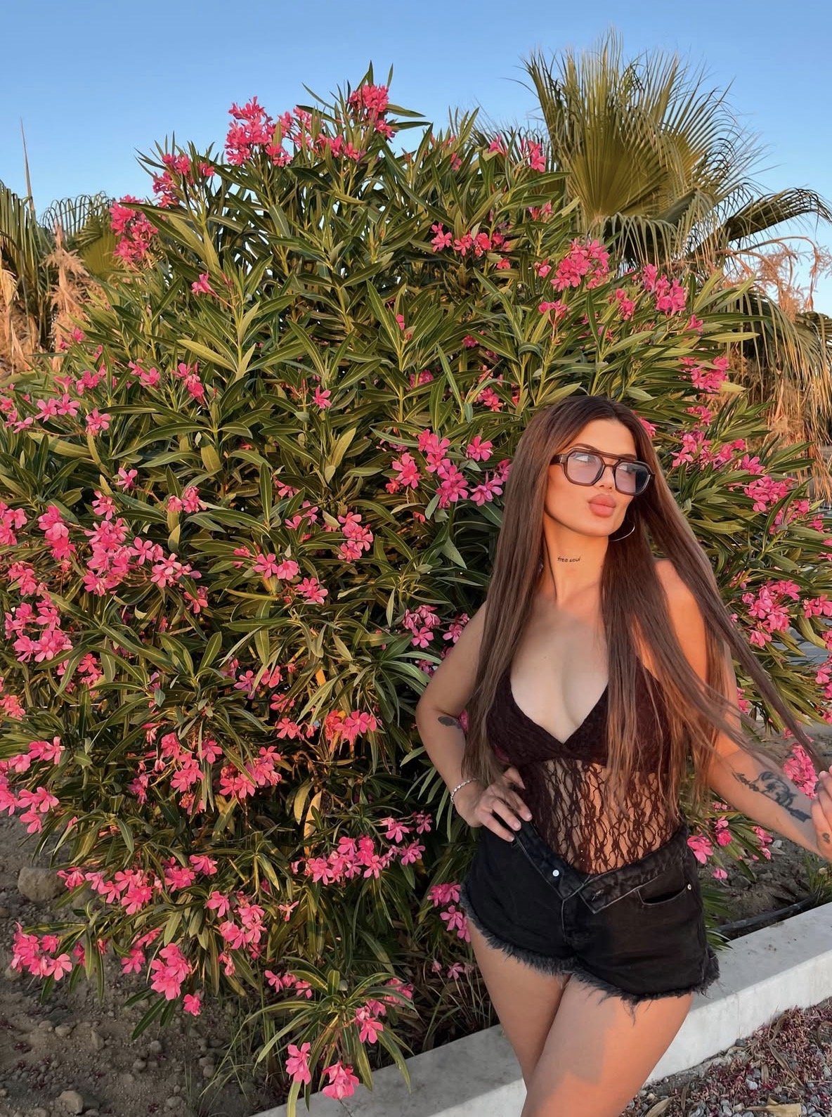 Woman with long hair and sunglasses posing beside a pink flowering shrub in sunny outdoor setting, wearing a black lace top and shorts.