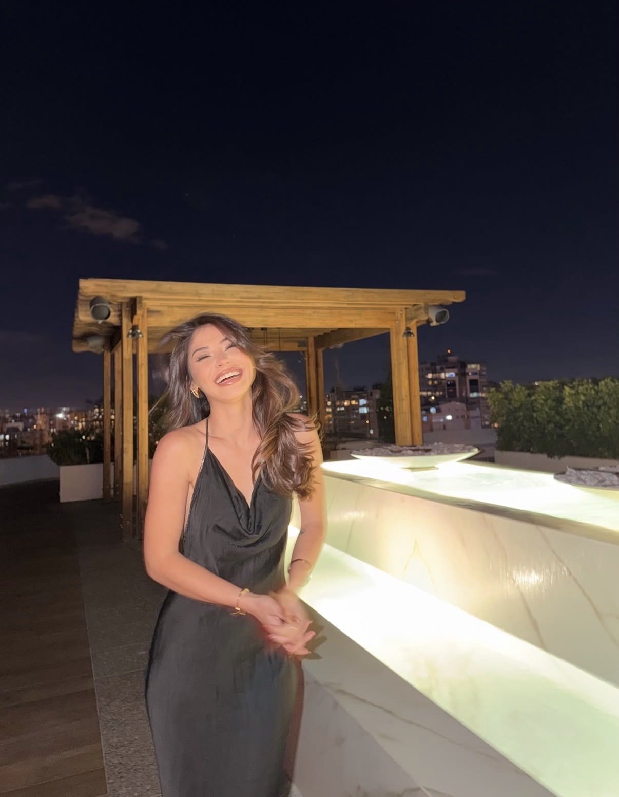 Woman in a black halter dress laughing on a rooftop at night near a glowing white railing and wooden canopy.",