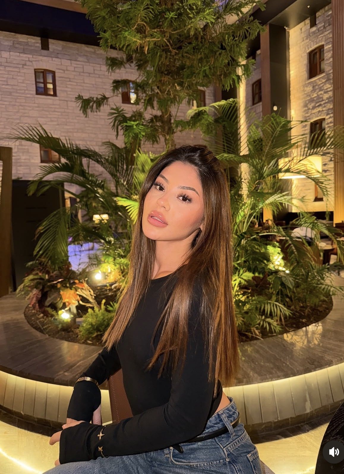 Portrait of a young woman with long brown hair in a black top, posing in an indoor tropical garden.