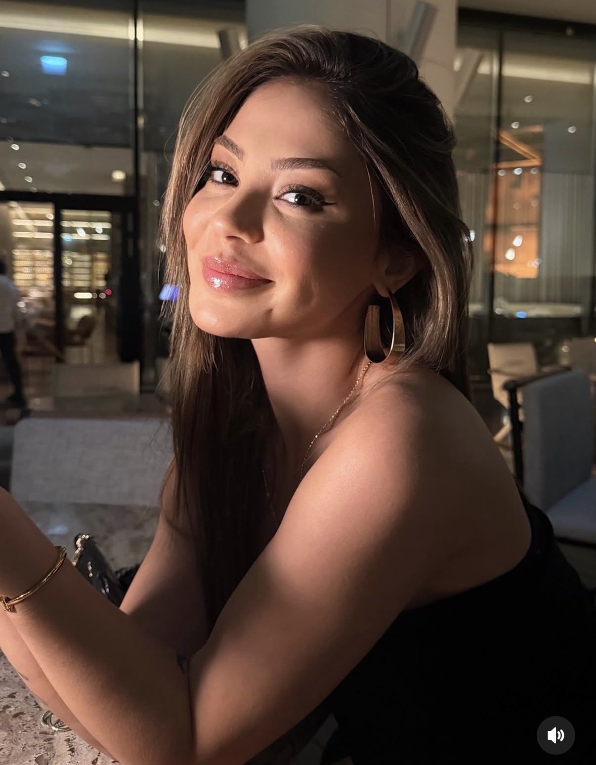 Smiling woman with long hair and hoop earrings at a stylish indoor lounge, warm lighting in background.
