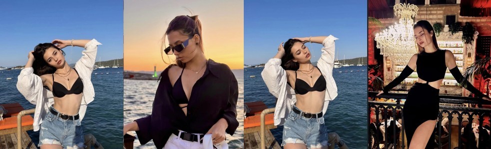 Woman posing on a waterfront boardwalk in a black bralette, denim shorts, and a white shirt, against the sea and boats.