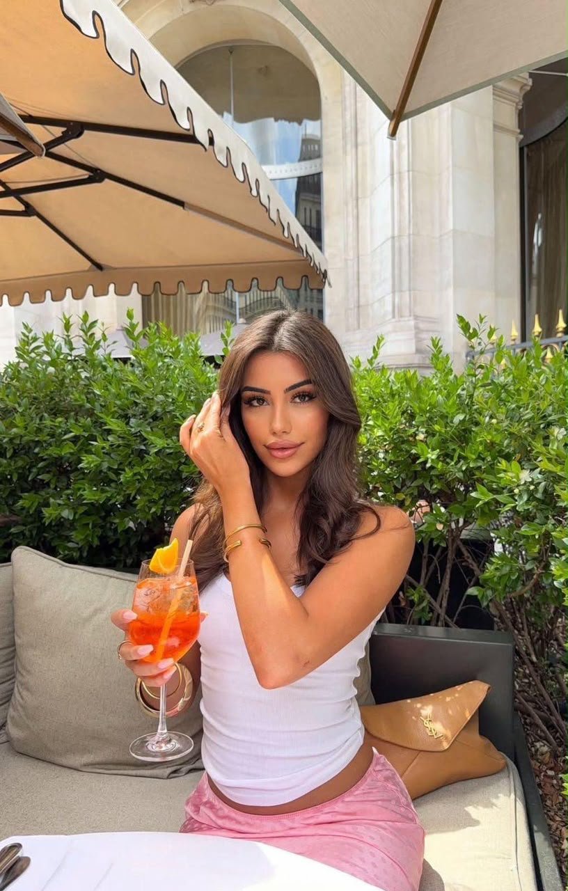 Woman sitting at an outdoor cafe table, wearing a white tank top and pink shorts, holding an orange cocktail with an orange slice and straw while smiling at the camera.