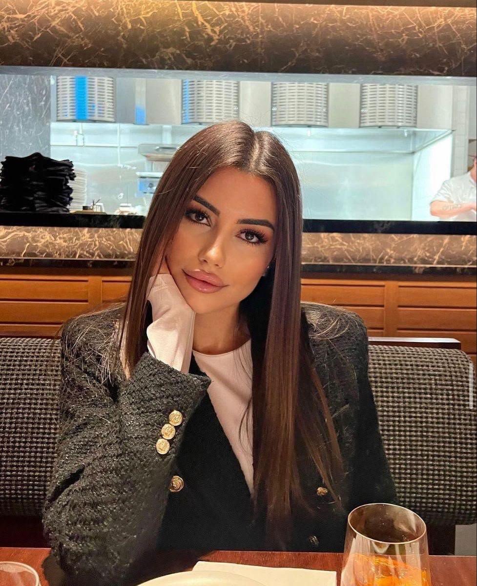 Woman with long brown hair sits at a restaurant table, resting her chin on her hand, wearing a dark blazer with gold buttons; open kitchen behind her.