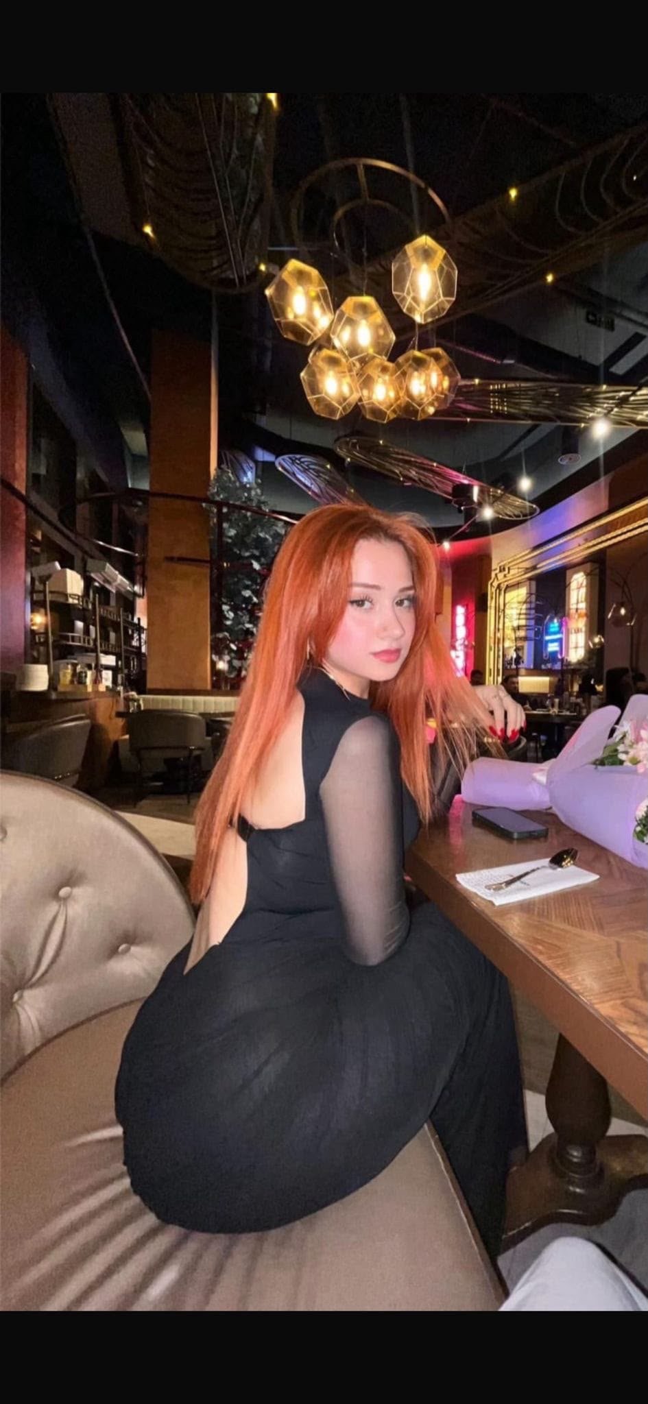 Woman with long orange hair wearing a backless black dress sits at a table in a dimly lit restaurant, turning to look at the camera.
