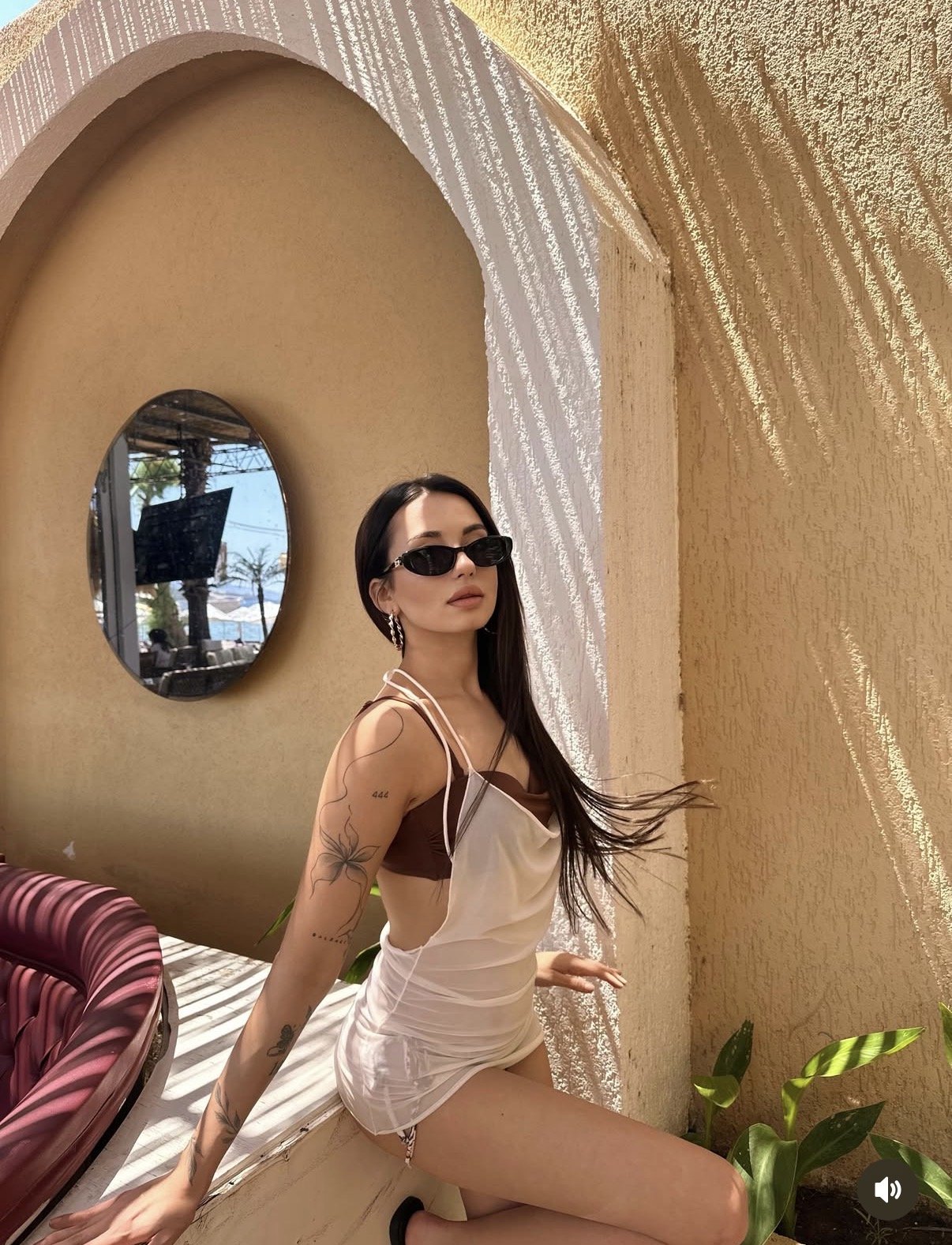 Woman in a white swimsuit with a brown bikini top posing beside a curved arch and round mirror, wearing sunglasses outdoors.