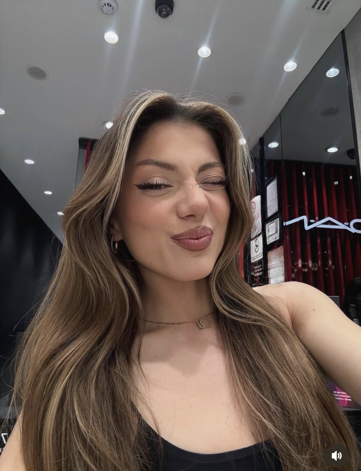 Smiling woman taking a selfie in a cosmetics store, MAC logo visible in the background, long wavy hair.