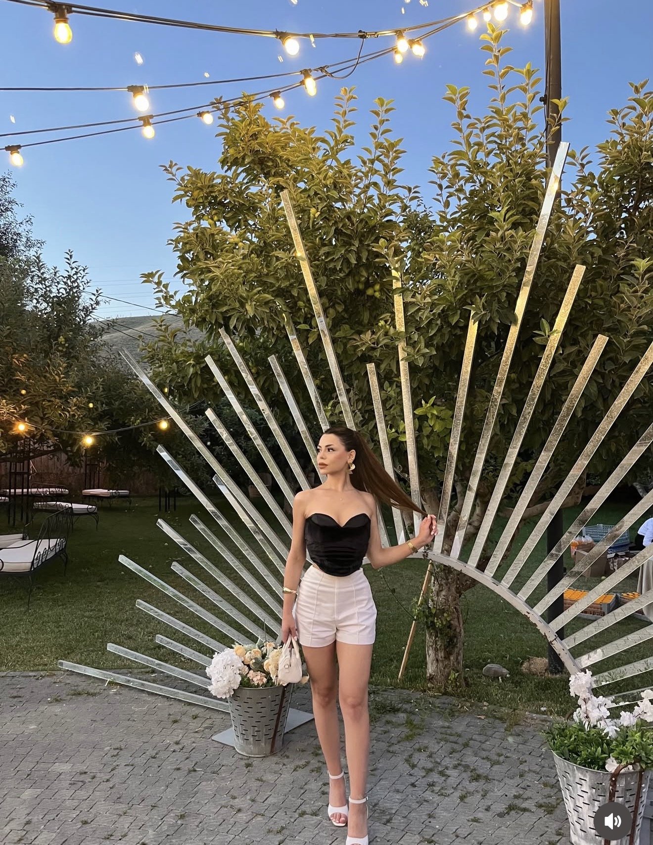 Woman in black bustier top and white shorts posing outdoors beside a metal fan sculpture and string lights