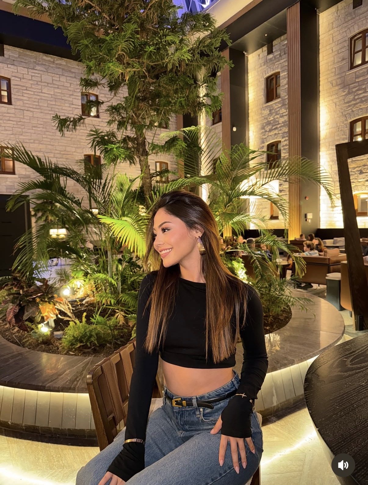 Smiling woman in a black crop top and jeans sits in a lush indoor atrium with palm plants and warm lighting.