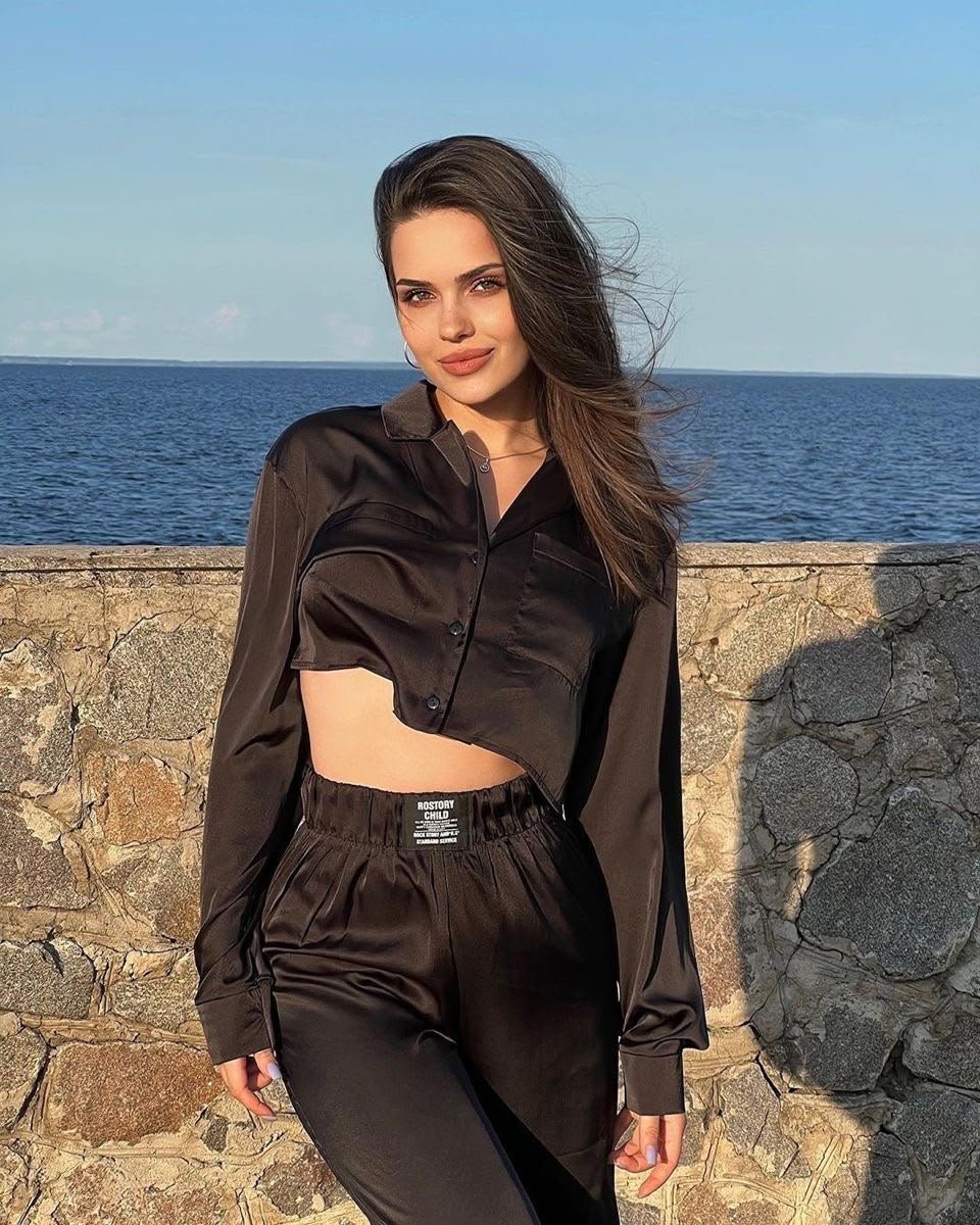 Woman posing by a seaside stone wall in a black cropped satin top and matching pants, sunlit with the ocean behind her.