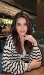 Woman with long dark hair wearing a black-and-white zigzag knit cardigan sits at a wooden table in a cafe, hand resting on her chin.
