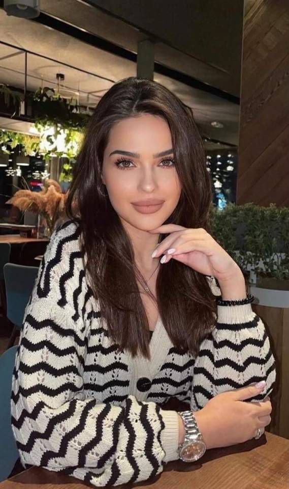 Woman with long dark hair wearing a black-and-white zigzag knit cardigan sits at a wooden table in a cafe, hand resting on her chin.