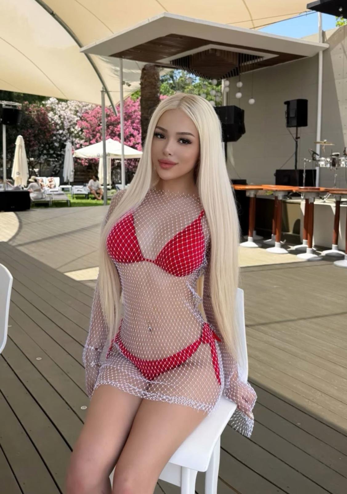 Young woman with long blonde hair in a red bikini under a white fishnet cover, seated at an outdoor event with a stage behind.