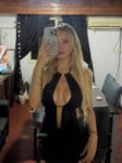 Woman with long blonde hair takes a mirror selfie in a black halter dress with a plunging neckline in a living room setting.