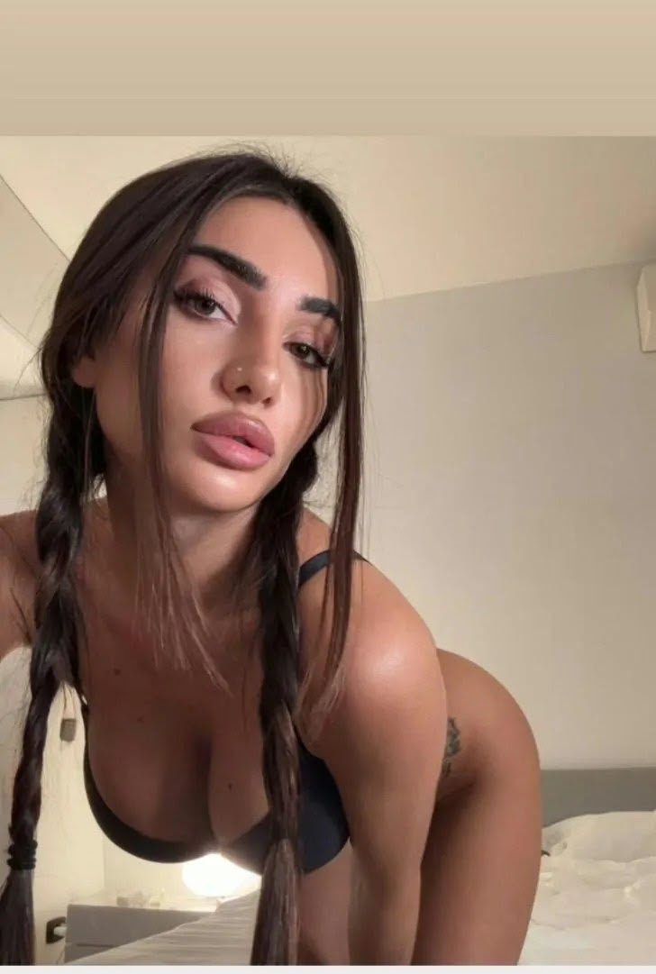 Young woman with two braids wearing a black bra, leaning toward the camera in a bedroom.