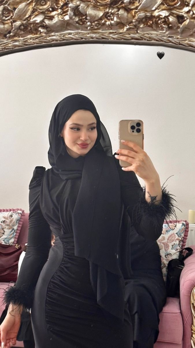 Woman in a black hijab and elegant dress taking a mirror selfie in a room with pink seating and a gold ornate frame above the mirror