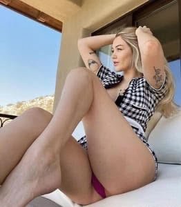 Woman sunbathing on a balcony or patio in a black-and-white plaid shirt and pink bikini bottom, arms resting behind her head.