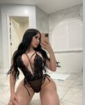Woman in black lace lingerie posing on a marble bathroom floor, taking a mirror selfie with a gold iPhone.