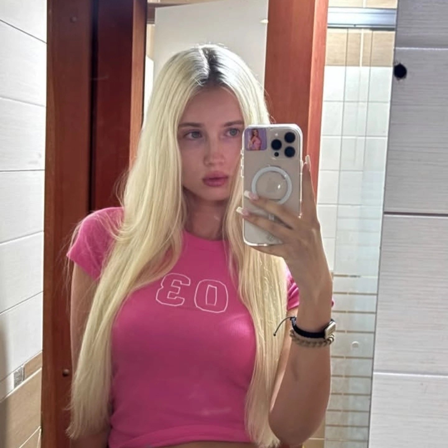 Young woman with long blonde hair taking a mirror selfie in a bathroom, wearing a pink T-shirt and holding a phone with a sticker on the back.